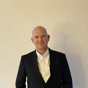 Ein Mann in schwarzem Anzug und weißem Hemd steht lächelnd vor einer schlichten beigen Wand.