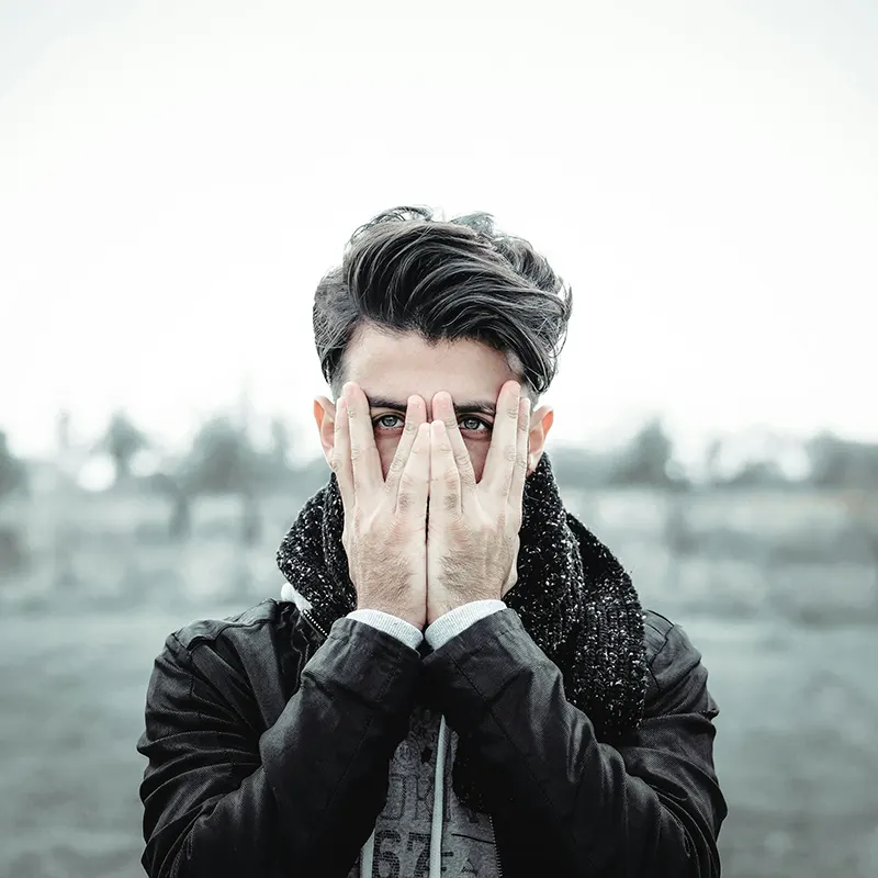 Ein Mann mit dunklem Haar bedeckt sein Gesicht mit den Händen und blickt durch seine Finger hindurch, draußen an einem nebligen Tag.